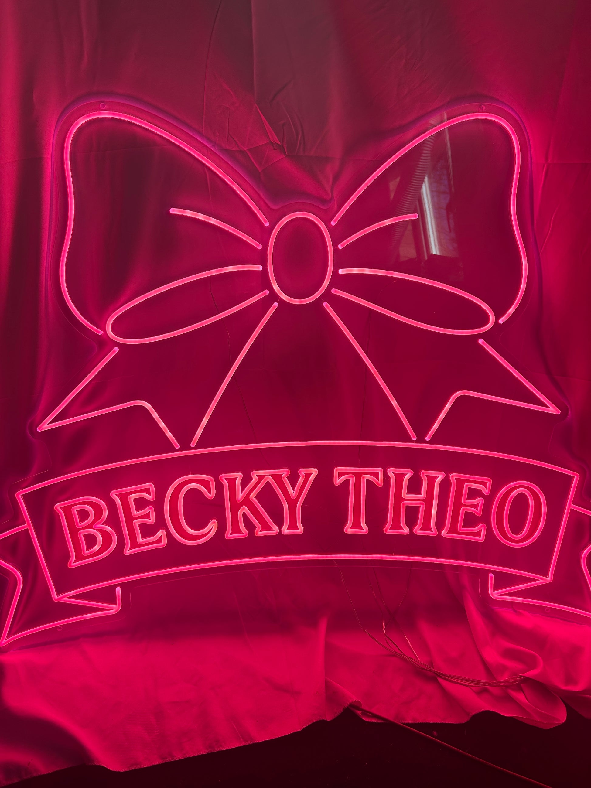 Neon sign with a bow design and 'BECKY THEO' text on a dark background