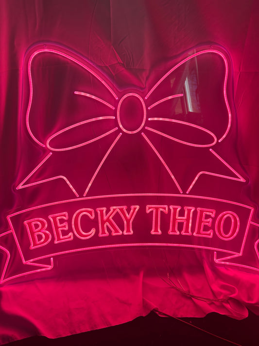 Neon sign with a bow design and 'BECKY THEO' text on a dark background
